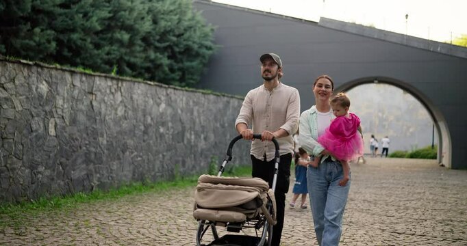 Happy brunette woman walking with her husband who is pushing stroller while the girl in a pink dress is sitting on her mother lap during a walk on the cobblestones in the park