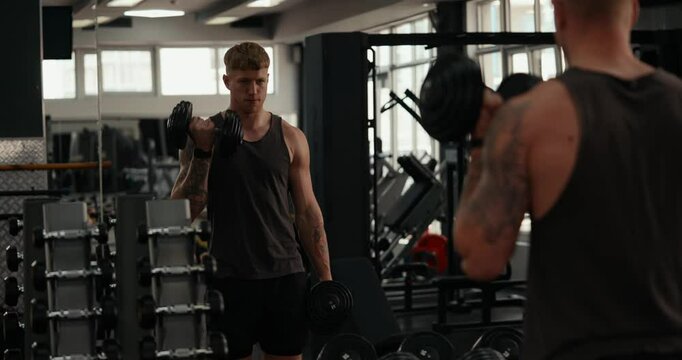Confident guy athlete fitness trainer in a gray T-shirt performs alternate dumbbell presses to his shoulders during a diligent fitness class in a modern gym