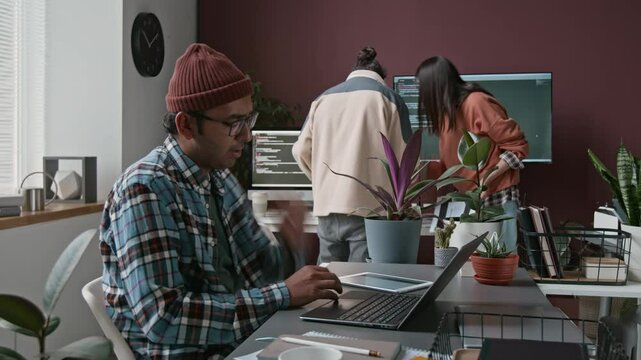 Medium full timelapse of young Indian male programmer in checked shirt and beanie arriving at work, sitting at desk, writing code on laptop and tablet, thinking, stretching, colleagues in background