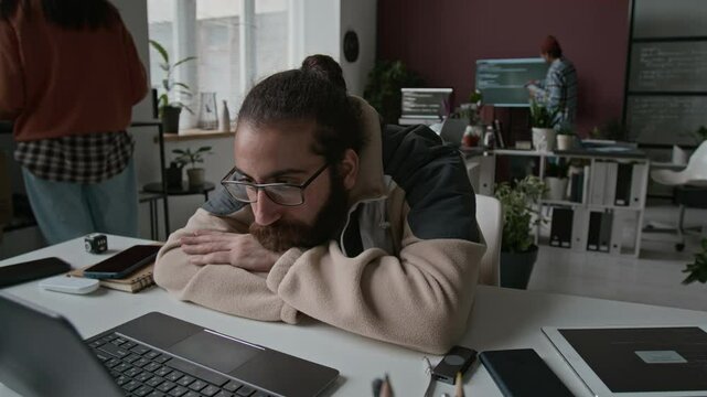 Medium time lapse shot of Middle Eastern male software developer lying on desk at work in busy office, staring at laptop screen with code with apathy and despair, stressed about approaching deadline