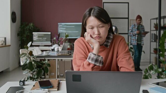 Medium timelapse shot of sleepy, burnt-out young Asian female IT engineer sitting at desk at work in busy office, staring at laptop screen with code with apathy, unable to fix bug in code