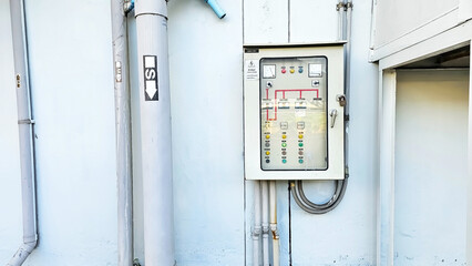 A control panel mounted on a wall with various switches and indicators, alongside a vertical pipe, showcasing industrial elements in a minimalistic environment.