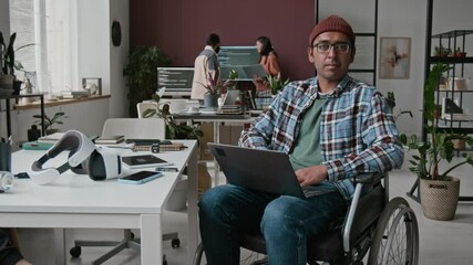 Medium timelapse portrait of Indian male software developer in glasses, beanie sitting in wheelchair in IT startup office, with laptop, looking at camera, colleagues working on project in background - Powered by Adobe