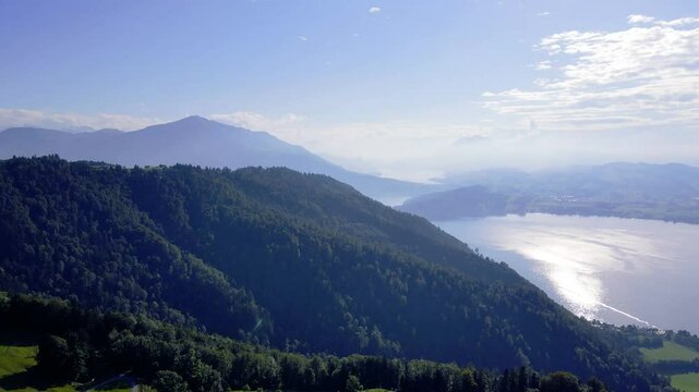 A spectacular drone shot showcasing Switzerland's lush green landscape featuring Lake Zug and Lake Lucerne with Mount Rigi and Mount Pilatus in the background under a hazy atmosphere