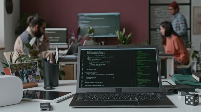 Timelapse of laptop computer on desk in IT startup, lines of code executed on black screen, virtual reality headset on desk. Diverse team collaborating on project in background, discussing algorithms