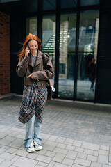 Full length vertical portrait of attractive young woman with flowing red hair enjoying cheerful phone conversation standing on city street on summer day. Smiling redhead female contemporary lifestyle