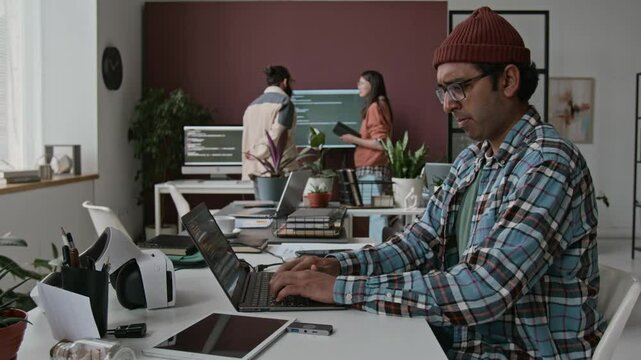 Medium timelapse of Indian male software engineer in checked shirt, beanie sitting at desk in corporate office, writing computer code on laptop, colleagues discussing IT project in background