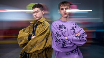 Obraz premium Two young individuals stand confidently against a blurred urban background with streaks of light, showcasing contrasting street fashion. 