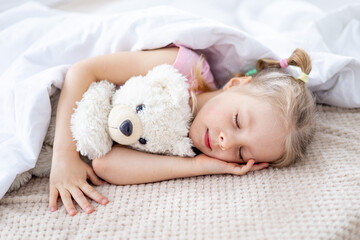 a blonde baby girl is sleeping sweetly under a white blanket holding a teddy bear in her hands on a white bed in a bright bedroom