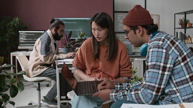 Medium shot of Asian female product manager and Indian male website designer sitting in office, studying software prototype on laptop screen and discussing improvements for better user experience