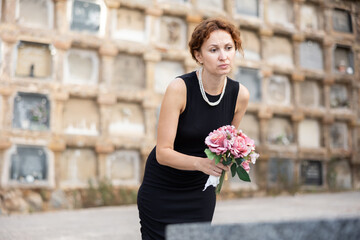 Fototapeta premium Woman in mourning wearing black dress gently resting pink bouquet on grave with sorrowful expression showing quiet reflection and deep loss in cemetery setting