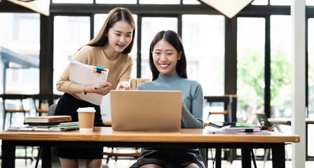 Two attractive asian business women using laptop, standing and talking about analyzing documents in workplace office