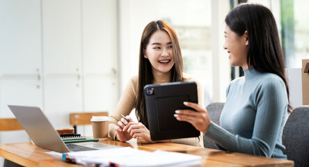 Two young asian business women sitting using laptop and talking about analyzing documents in workplace office