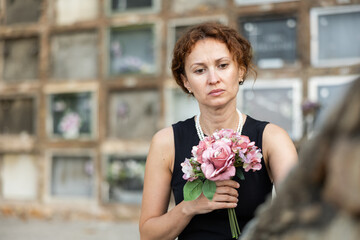Fototapeta premium Middle aged woman wearing black dress and pearls holding bouquet of pink flowers, gazing sorrowfully at grave, reflecting deeply on loss in quiet cemetery
