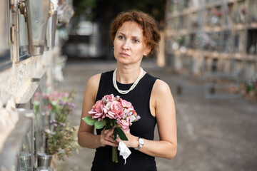 Elegant grieving woman in forties, wearing formal black dress and pearls, standing with bouquet in...