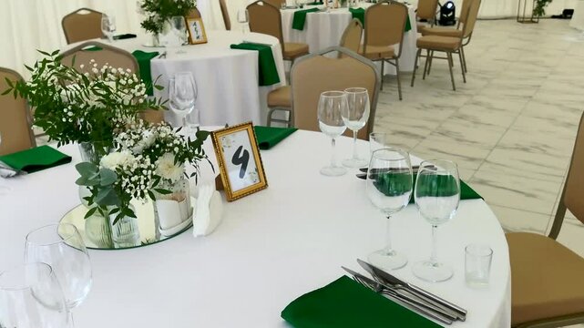A snow-white wedding tent with round tables covered with white tablecloths, green napkins and glasses for drinks, lighting devices are working. A table set for a wedding reception under a tent.