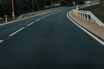 A smooth, empty road winds through a wooded area with dense trees on one side and open fields on the other. The soft evening light creates a peaceful atmosphere with ample copy space in the sky and on