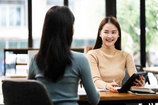 Two asian business women sitting at desk using tablet and talking about analyzing documents in workplace office