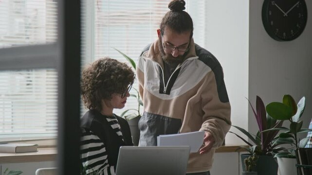 Medium shot of young Caucasian female manager in glasses sitting on desk, talking to Middle Eastern male team member, discussing documents with presentation or data
