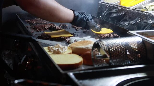 Video of a hand placing cheese on top of cooking smash burgers on a hot griddle, melting the cheese over the hamburgers in a professional kitchen setting