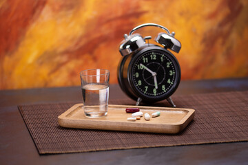 Pills, water glass, and alarm clock on brown mat surface