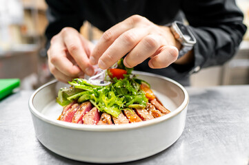 A close-up of hands garnishing a fresh salad with sliced onions, served on a plate with grilled meat and greens. The vibrant colors and textures highlight a delicious and healthy meal