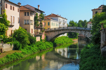 Fototapeta premium Charming view of a historic stone bridge over a tranquil canal in Vicenza, Italy. Colorful buildings and lush greenery line the waterway, creating a idyllic Italian cityscape