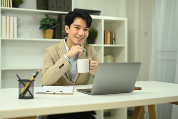 Portrait of confident young businessman holding cup of coffee sitting at his workplace