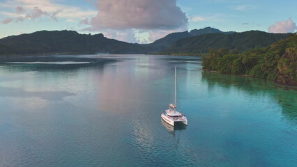 Aerial view catamaran sailing calm turquoise water lagoon near tropical island with lush green mountains and a beautiful sunset in background. Remote wild nature paradise, exotic summer luxury travel