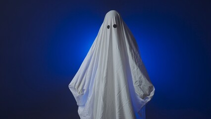 Funny Halloween ghost in white sheet makes scary gestures on background with blue spotlight in studio. Happy Halloween spooky concept.