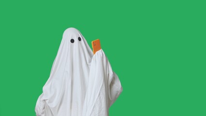 Funny halloween ghost in white sheet talking on smartphone by video call, isolated on chroma key...
