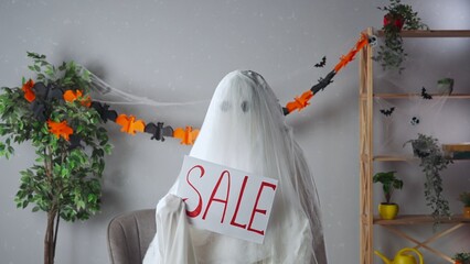 Funny halloween ghost in white sheet with black eyes in bright room holding showing paper sign with...