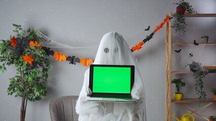 Funny halloween ghost in white sheet with black eyes in bright room holding laptop with chroma key...