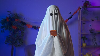 Funny halloween ghost in white sheet with black eyes in dark room texting on smartphone, sending message in app. Happy halloween spooky concept.