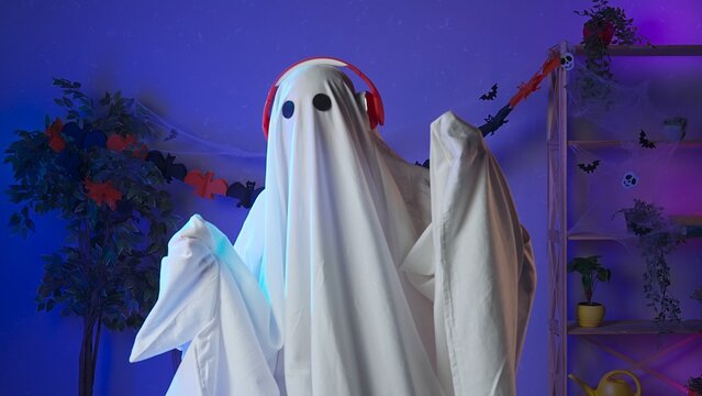 Funny halloween ghost in white sheet with black eyes in dark room dancing and listening music in headphones. Happy halloween spooky concept.