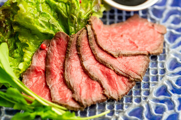 青いガラス皿に美しく並べられた、鮮やかな赤身が目を引くローストビーフ。しっとりときめ細やかな地鶏のような質感と、絶妙な厚みのスライスが食欲をそそる一品。添えられたレタスの緑やピンクペッパーの赤が彩りを添える、パーティーやディナーの主役に最適なクローズアップ素材。 | Sliced roast beef with lettuce and pink peppers on a blue plate.