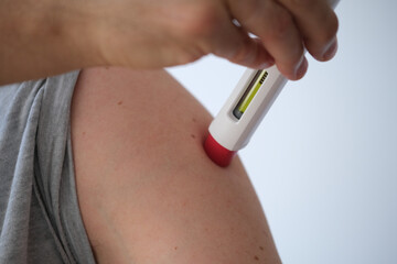 an anonymous person injecting himself a treatment such as insulin or Adalimumab, Etanercept or epinephrine into his shoulder. It is a white pen injector with red head for health care