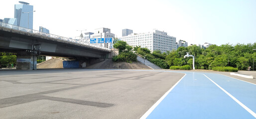 blue running track, on a city park, in Seoul, a paved area with a blue-painted section, surrounded by lush green trees and bushes. buildings and structures, on a sunny day with a clear sky