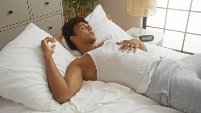 Young man with curly hair relaxing in bed wearing white tank top and gray pants in a cozy bedroom with white bedding and a dresser in the background