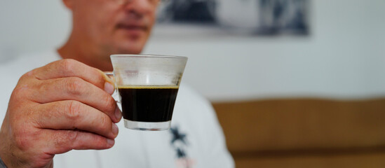 hombre con la cara desenfocada levantando un a taza transparente de café ,con el fondo gris y marron