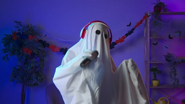 Funny halloween ghost in white sheet with black eyes in dark room dancing to music in headphones sings in karaoke microphone. Happy halloween spooky concept.