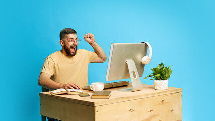 Young emotional bearded man sitting at desk and interacting with computer, expressing excitement...