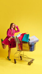 Young woman in stylish bright dress carrying shopping cart with many multicolored shopping bags inside against yellow studio background. Concept of shopping, sales, Black Friday, Cyber Monday