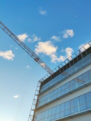 Fototapeta premium In this image, a sleek, glass-structured building is juxtaposed with a towering crane against a bright blue sky.