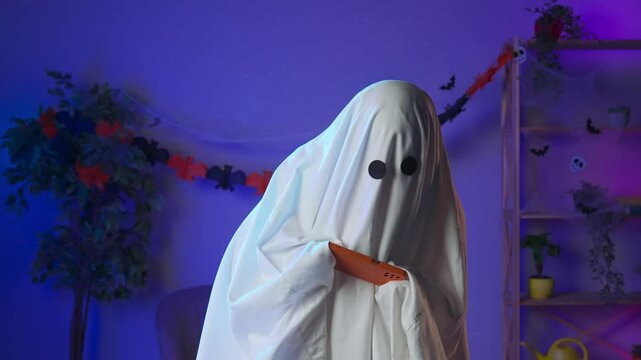 Funny halloween ghost in white sheet with black eyes in dark room playing video online game on smartphone. Happy halloween spooky concept.