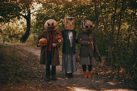 Three strange people in terrifying masks and odd clothes standing in autumn woods, creating creepy Halloween mystery. Concept of Halloween, spooky season, retro style.