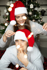 Young cheerful couple man and woman home pajamas and red Santa hats near Christmas tree with lights.