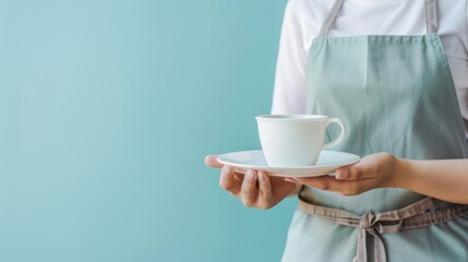 A person wearing a light green apron holds a cup and saucer, presenting a minimalist, welcoming scene against a teal background.