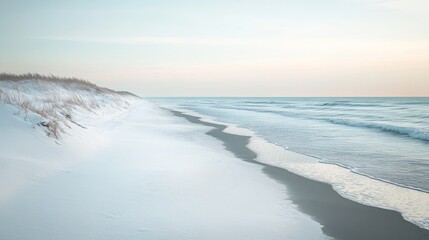 Obraz premium A calm winter beach with snow on the dunes and icy waves lapping at the shore, under a pale blue sky
