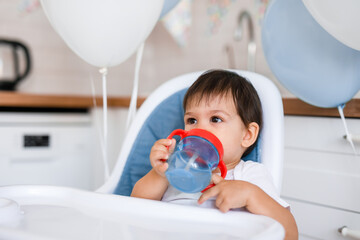 Little baby boy sitting in high chair at home on white kitchen and drinking water from sippy cup on background with balloons.